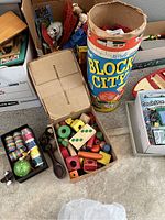 Photo showing vintage painted wooden blocks, puzzle pieces, and colorful wooden toy perforated pieces in a cardboard box. Large cylinder labeled 'Block City' containing plastic blocks visible in background.