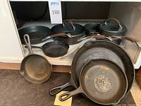 Overview of four cast iron skillets and Circulon cookware including pots, pans, and a large glass lid on a shelf.