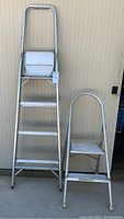 Photo shows a taller aluminum 3-step ladder next to a smaller 2-step aluminum stool with a top handle, both against a beige wall.