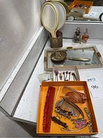 Vanity accessories displayed on a mirrored surface with two small perfume or cologne bottles, a brass or bronze-toned ornate hand mirror and brush, and a mirrored tray.