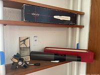 Spotting scope, mounting hardware, original box and pamphlet shown on wooden shelf.