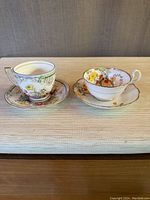 Two antique floral teacups with matching saucers side by side on a beige textured fabric. Melba cup on left with green and floral accents, Bell cup on right with scalloped edge and floral design.
