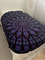 Full view of the circular crochet afghan on a bed, showing the intricate stained glass window inspired design with predominantly purple, blue, and black colors.