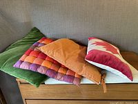 Four cushions stacked horizontally on a wooden surface showing different fabrics and colors: velvet green, quilted silk orange/red, burnt orange cotton, and Canada flag cushion.