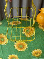 Yellow wire metal carrier for insulated carafe, rectangular base with handle, shown on sunflower patterned tablecloth.