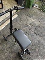Black knee scooter on outdoor asphalt surface showing padded knee rest, handlebar, and frame.
