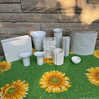 Collection of eleven white ceramic vases arranged on a green sunflower tablecloth in front of stone wall backdrop. Various shapes including rectangular, cylindrical, and small bud vases with different embossed and textured details.