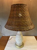 Full view of the lamp showing the wicker shade and pottery base with metal stand on a wooden surface, lamp turned on.