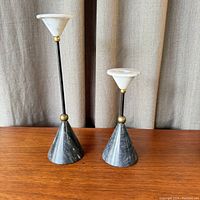 Pair of two Postmodern Regency style candleholders made of dark marble bases with white markings, black metal rods, gold accents, and white marble candle holders on top shown on wooden surface with light curtain background.