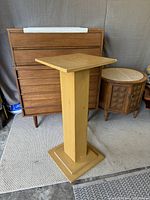 Full view of the wooden pedestal standing on a carpeted floor near other wooden furniture pieces.