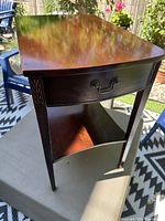 Front angle photo showing the dark brown wooden side table with one drawer and lower shelf, featuring carved legs and metal handle.