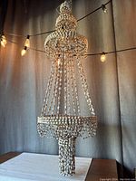 Full view of the large vintage hanging cowry shell planter chandelier suspended in front of curtains, showcasing its tiered shell strands and kettle silhouette.