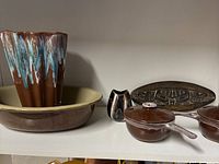 Lot of seven items: Colloma Pottery drip glaze vase set in beige bowl, brown ceramic casseroles, small creamer pitcher, and oval serving platter on shelf.