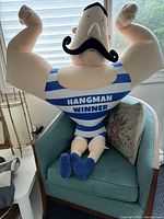Front view of the XL stuffed muscle man toy sitting on a blue chair with 'HANGMAN WINNER' on chest and large flexed arms.