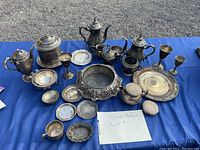 Full view of assorted antique silver and silver plated items arranged on blue cloth, including pitchers, kettles, goblets, trays, and bowls all with tarnished patina.