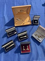 Photo showing multiple assorted cufflinks and tie clips placed on blue fabric, each in their original boxes from brands like Ambassador and Imperial.