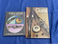 Photo showing the two books: 'Firearms' by Howard Ricketts and 'The Pleasure of Guns' by Joseph G Rose and Robin May, placed side-by-side on a blue fabric background.