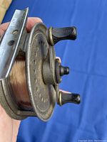 Close-up of vintage metal fly fishing reel showing the two black handles and perforated circular frame with fishing line.