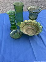 Five assorted green glassware pieces including bowls and vases on a blue cloth surface