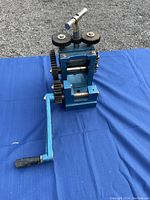 Photo showing blue manual rolling mill placed on blue cloth with visible gears and crank handle.