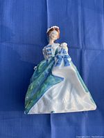 Porcelain figurine of a woman in a blue and green gown holding a bird, photographed against a blue background.