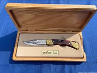 Photo of the Schrade special edition folding knife in an open wooden display box, showing the engraved blade and handle details.