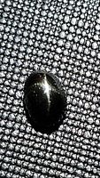 Close-up photo of the oval black star diopside gemstone showing the star effect on its smooth cabochon surface, placed on a textured black fabric background.