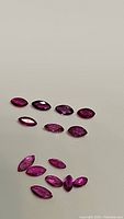 Several natural ruby gemstones in marquis cut, exhibiting vibrant exotic hues ranging from fuchsia, purple, red, to pink, laid out on a plain light background.