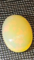 Oval cabochon cut fire opal showing translucent light body with vibrant orange and yellow internal flashes on dark textured background.