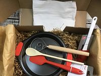 Box contents including 9-inch baking pan, wooden mixing wand and spoon, red silicone spatula, white silicone spatula with red handle, and white tea towel inside open cardboard box with shredded paper filler.