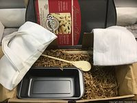 Box containing baking items including a 13-inch baking sheet pan, 8.5-inch baking pan, wooden spoon, wooden mixing wand, white tea towel, and small white tote bag, arranged on brown shredded packing material