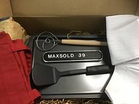 Opened box showing 13-inch baking sheet, wooden-handled mixing wand, black spatula, white tea towel, red apron, and small tote bag. Items neatly arranged and new.