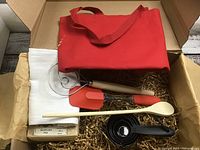 Photo showing red apron, wooden spoon, red-handled spatula, metal mixing wand, black measuring cups, and white tea towel in box