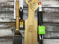 Top-down image showing a wooden-handled metal spatula, cleaning brush, bamboo wood scraper, and stainless steel tongs with black handles arranged horizontally on a rustic wood background.
