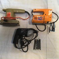 Black & Decker drill (black and gray) with attached power cord and multiple drill bits, Black & Decker 1/4 sheet sander in olive green with orange cord, and Mastercraft orange sabre saw with black handle and cord, with saw blades shown separately.