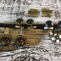 Front view of five pairs of cuff links and a set of shirt studs arranged on a wooden surface showing variation in design, color and shape.