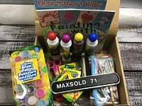 Photo shows a box filled with various kids art supplies, including packs of washable paints, Crayola crayons, several scissors, colored markers, and two books titled 'Heartsmart Kids Painting With'.