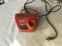 Milwaukee M12 battery charger with power cord visible, placed on a light-colored fabric surface, showing overall condition and vented design.