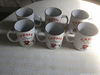 Six white ceramic mugs with 'CANADA' and cartoon moose playing hockey on front, arranged in two rows on a cloth surface.