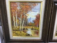 Framed landscape painting showing autumn birch trees next to a calm stream with fall foliage in orange and red hues.