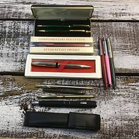 Overview of multiple pens laid flat on wooden surface showing boxed Parker pen, complimentary stylus pen set, three colored ballpoint pens, black pen with case, and additional metallic pens.