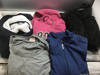Photo showing a collection of folded hoodies and tops including pink Roots hoodie, black fleece hoodie, grey Karen Scott top, blue zip-up, and more.
