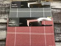 Two table runners stacked one on top of another showing grey and red color with white plaid lines, packaged with labels.