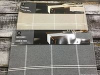 Photo showing two packaged table runners, one beige and one grey, stacked on a rustic wood surface.
