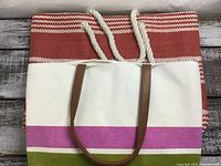 Photo of a white canvas tote bag with pink and green stripes on the bottom front panel and brown leather handles, placed in front of a larger red tote bag.