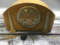 Front view of a rounded wooden mantel clock with glass-covered circular dial numbered 1 to 12 and two clock hands. Two accessories (a winding key and a pendulum) placed in front of the clock on wooden surface.