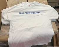 Stack of white Hanes T-shirts printed with 'First Class Returns' in blue on the front, folded and placed on a cardboard surface.