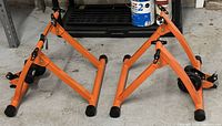 Two orange metal-framed bike trainers placed side by side on the floor, showing the structural framework and resistance units.