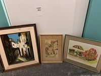 Three framed artworks on floor: an oil painting of a European street, a sketch-like watercolour, and a souvenir watercolour with flowers.