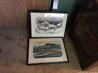 Two framed Asian prints leaning against a wooden wall, showing traditional landscape scenes with water and mountains